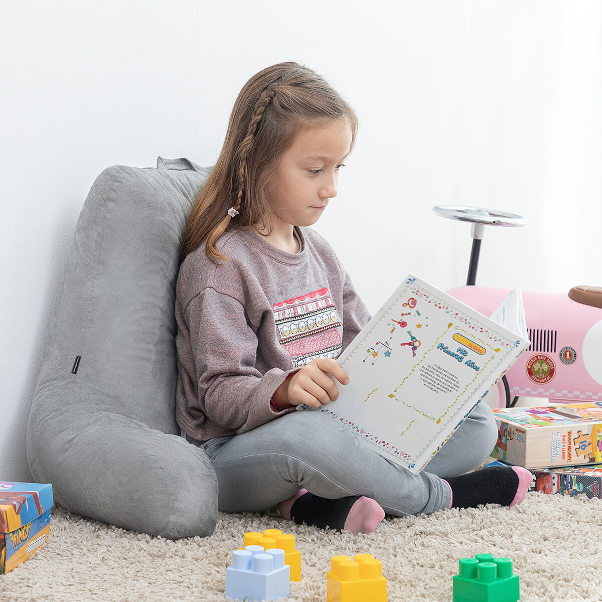 A child leaning against the grey reading pillow while reading a book on a soft carpet, showcasing its versatility for all ages.