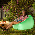 A woman is sitting on a glowing green inflatable armchair outdoors, holding a drink and a remote control. The setting is a garden with greenery and flowers, suggesting relaxation and leisure.