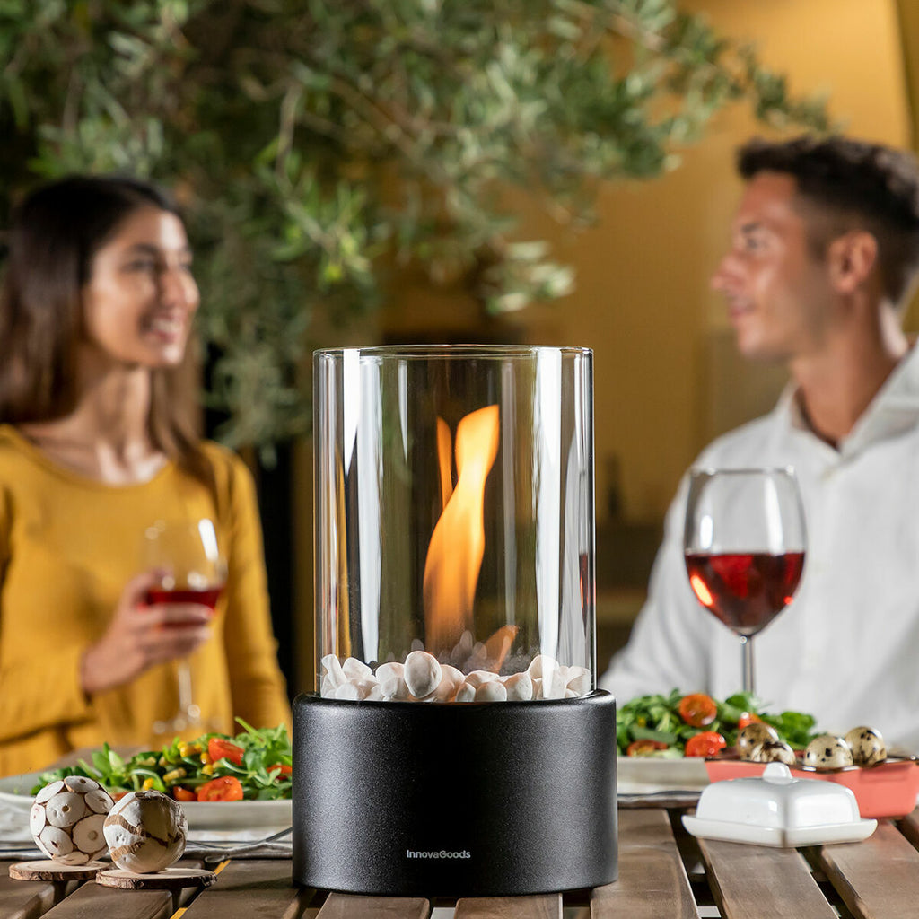 The tabletop fireplace is set on a wooden table, surrounded by a couple having a dinner with wine. The flame is visible inside the clear glass cylinder, which is adorned with white stones at the base. The setting is outdoors with a cozy, romantic ambiance.