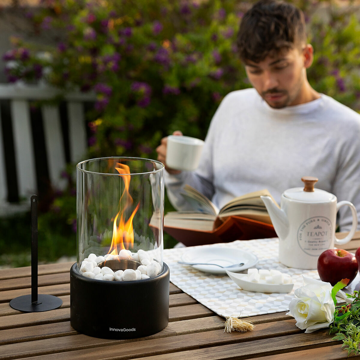 An outdoor setting with the fireplace on a wooden table next to a man reading a book and drinking tea, showcasing leisure time ambiance.