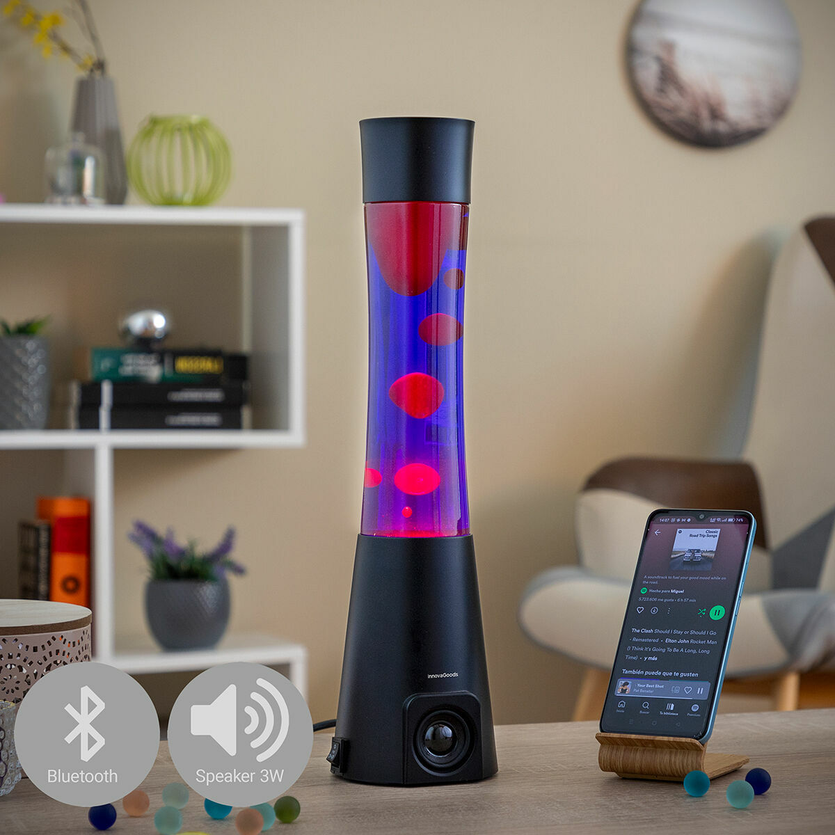 A lava lamp with vibrant red and blue liquid, set on a table with room decor in the background. A smartphone positioned next to it shows music playing, highlighting the Bluetooth feature.