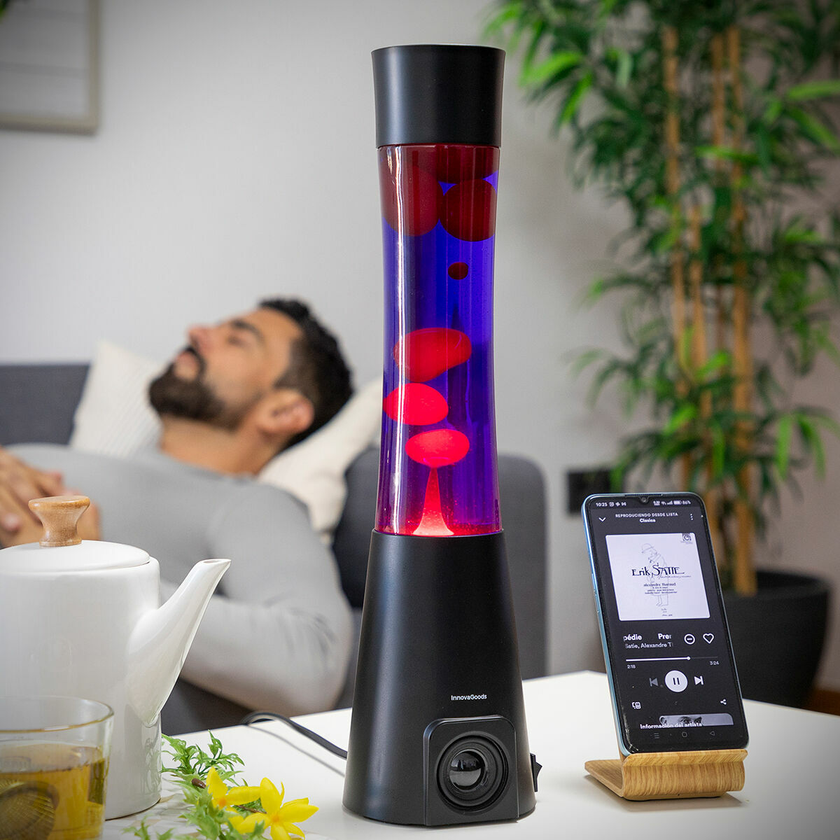 The lava lamp is in a relaxing setting with a man resting on a couch nearby and a smartphone is placed beside it, emphasizing its relaxing and technological features.