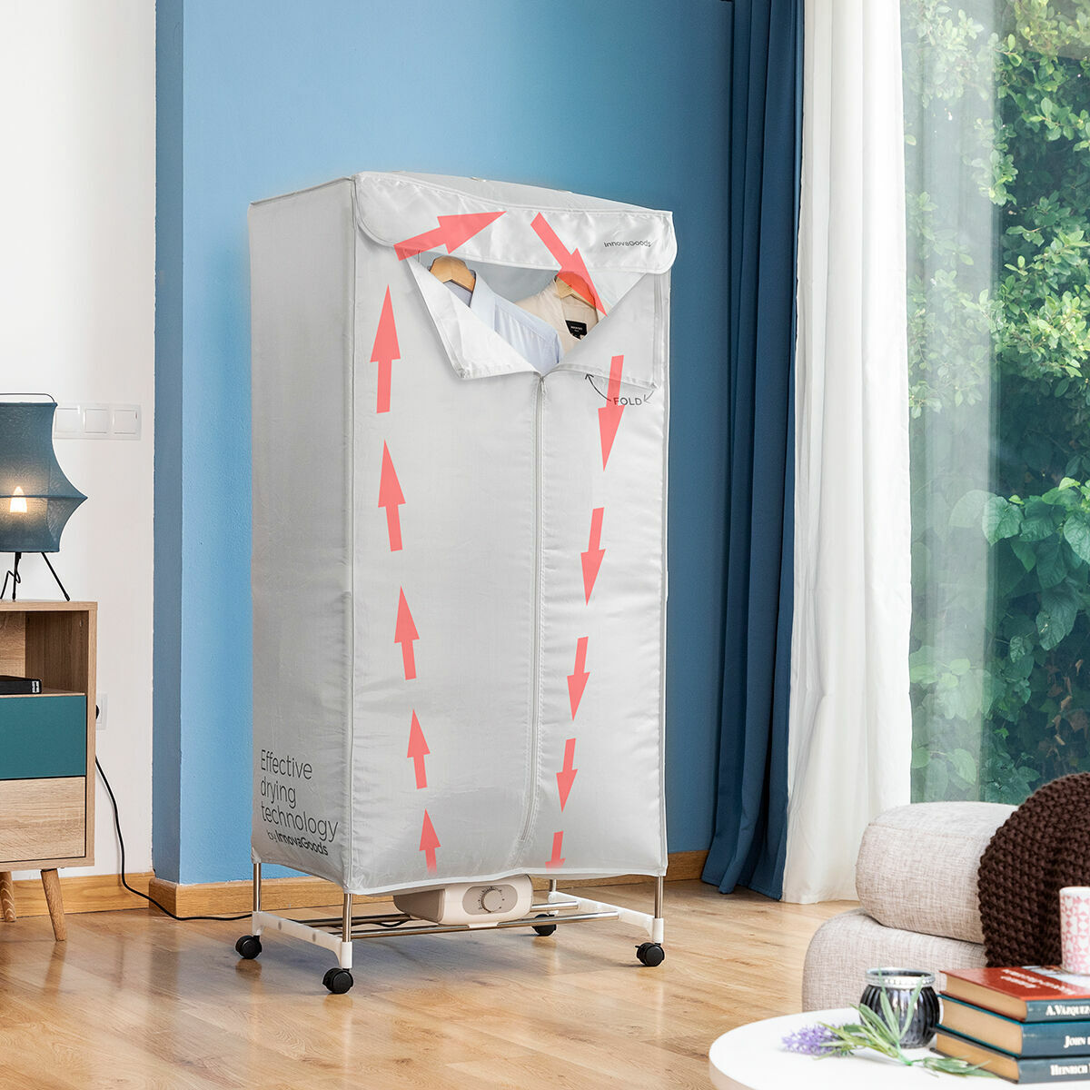 The InnovaGoods Portable Electric Dryer in a living room setting with wooden floors and blue walls. The silver fabric dryer cover is mounted on a frame with wheels, showcasing red arrows indicating air flow. It contains hangers with shirts visible through the top opening.