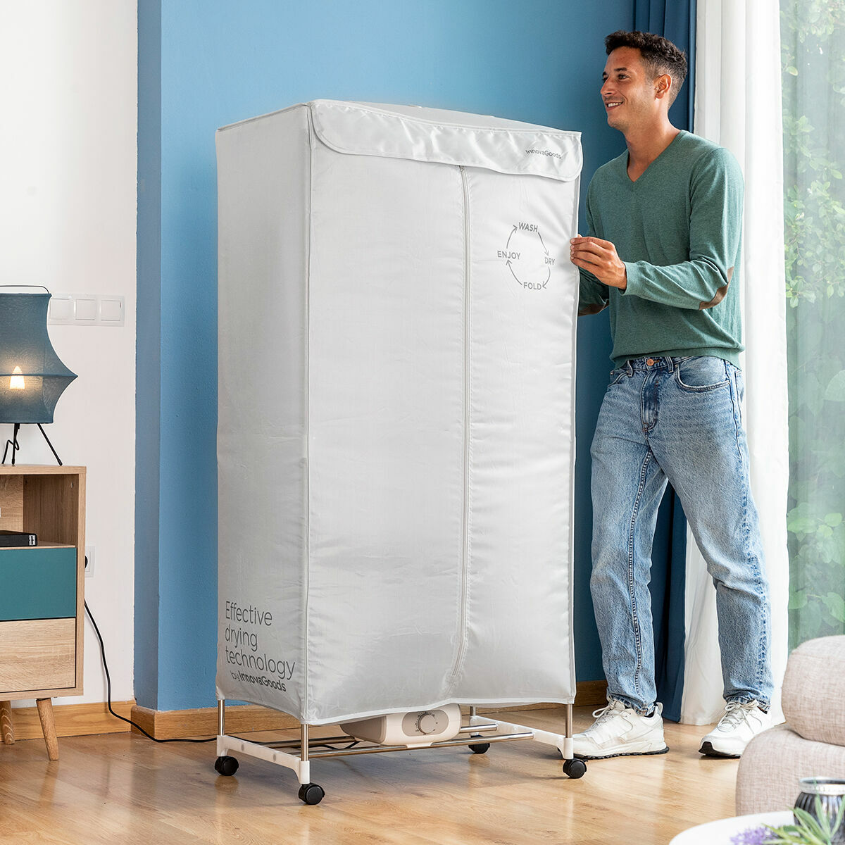 Person standing next to the assembled dryer in a living room setting. The silver cover is on, showcasing branding and a flow cycle graphic. Wooden floor and blue walls in the background.