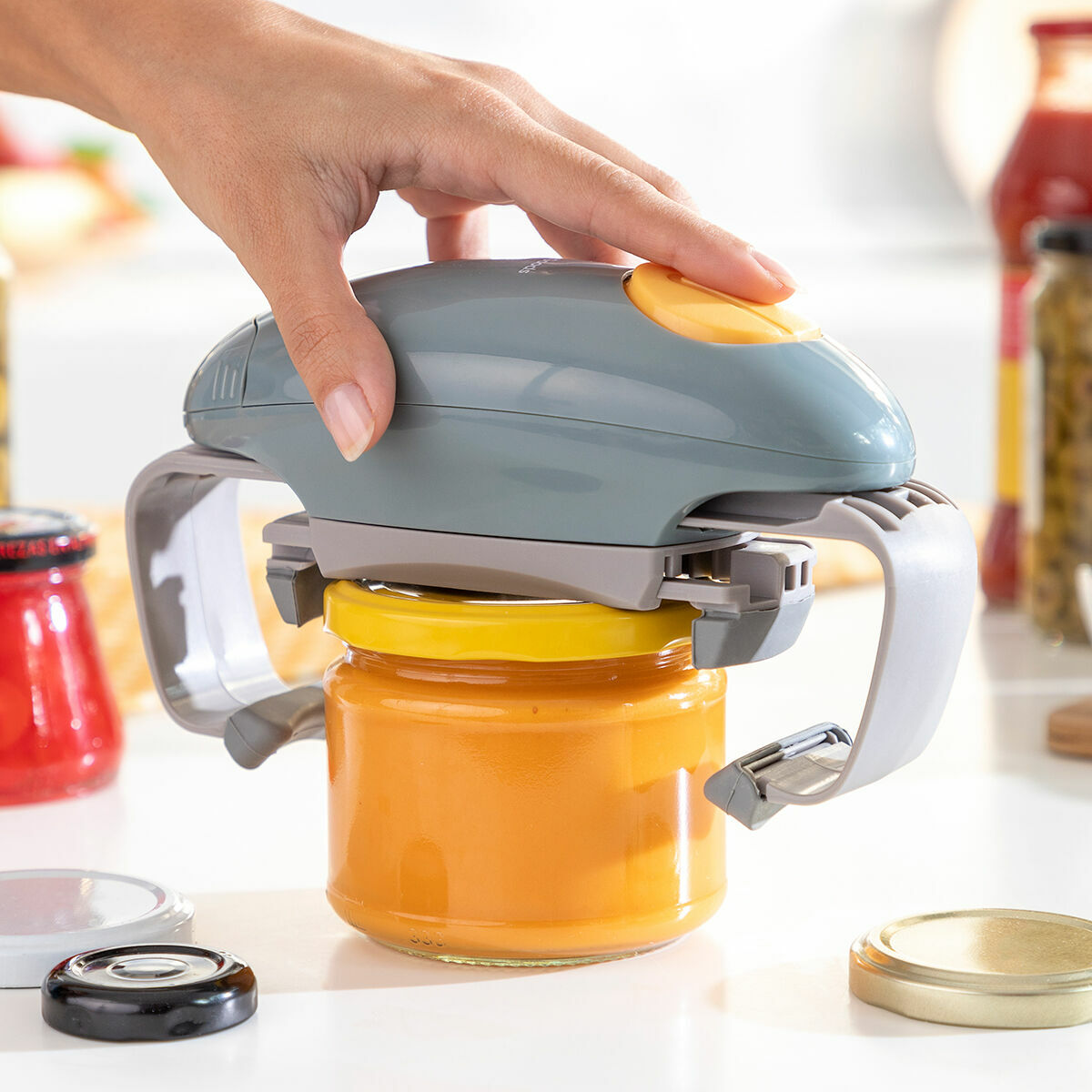A close-up image of the Electric Jar Opener Lloucan InnovaGoods in use. The opener is placed on a jar with orange contents, and a person's hand is pressing the yellow button, demonstrating its ergonomics. The setting is a kitchen with jars in the background.