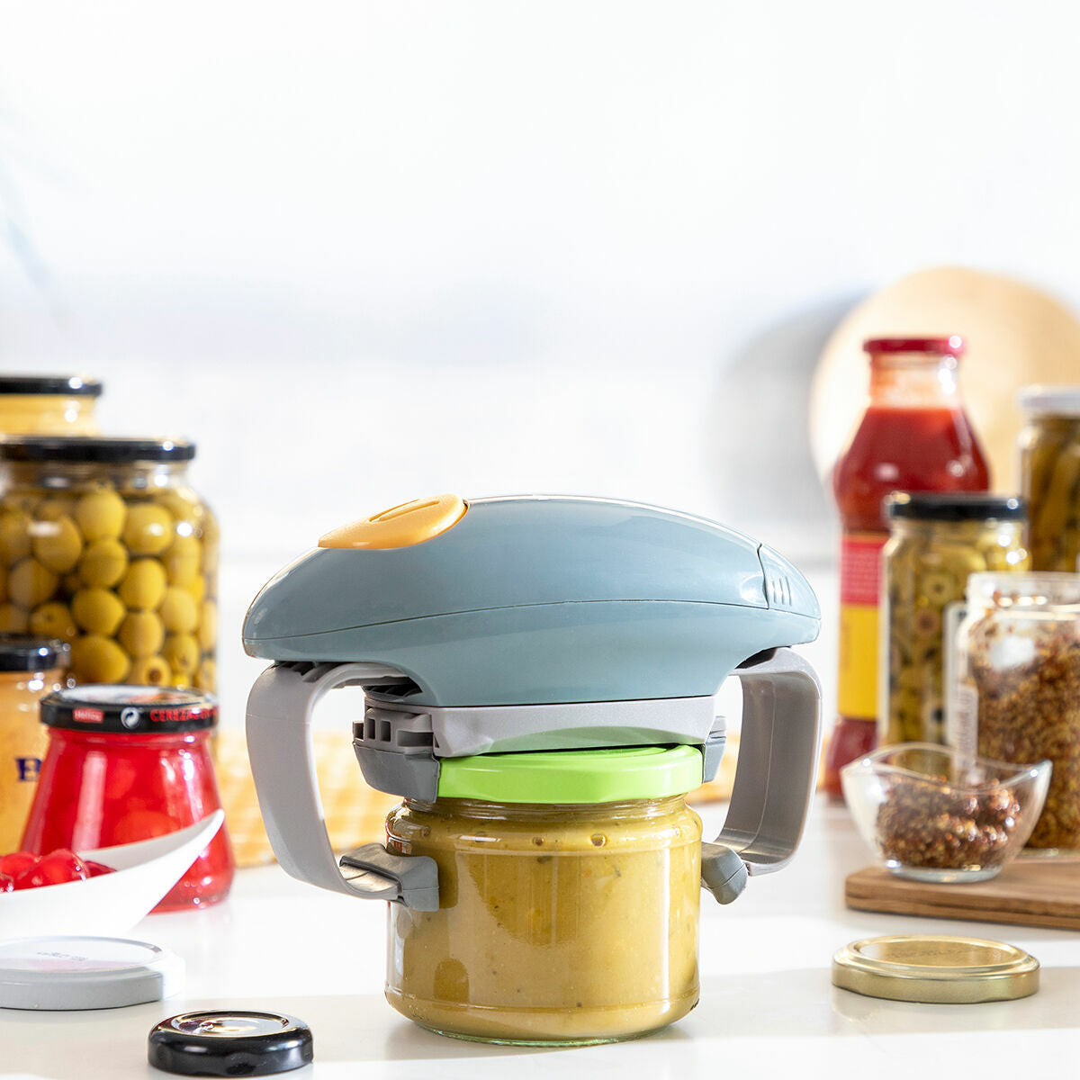 The Electric Jar Opener Lloucan InnovaGoods shown on a jar with a green lid, placed on a kitchen counter amidst other jars. This setting conveys ease of use in a practical environment.