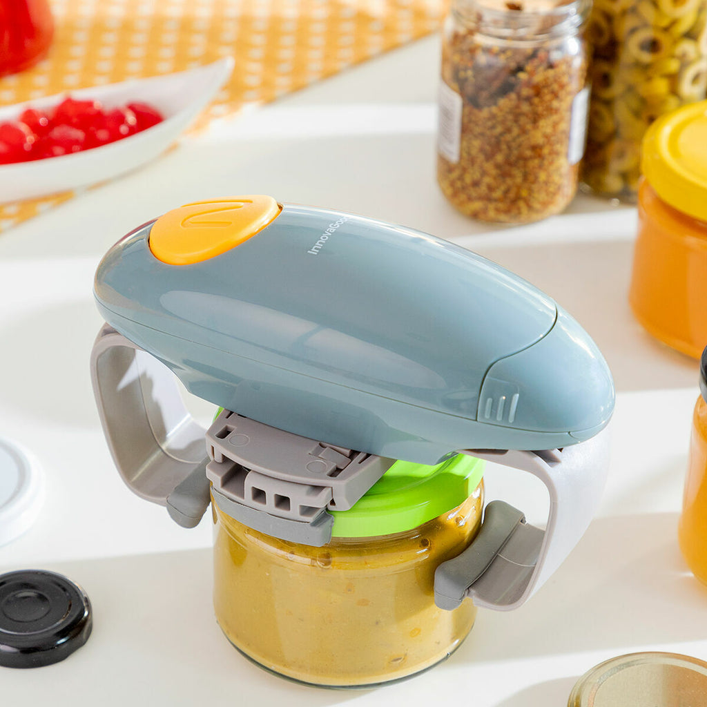 A top-down view of the Electric Jar Opener Lloucan InnovaGoods resting on a jar with a green lid. The blue body and yellow button are clearly visible, set against the backdrop of a kitchen.