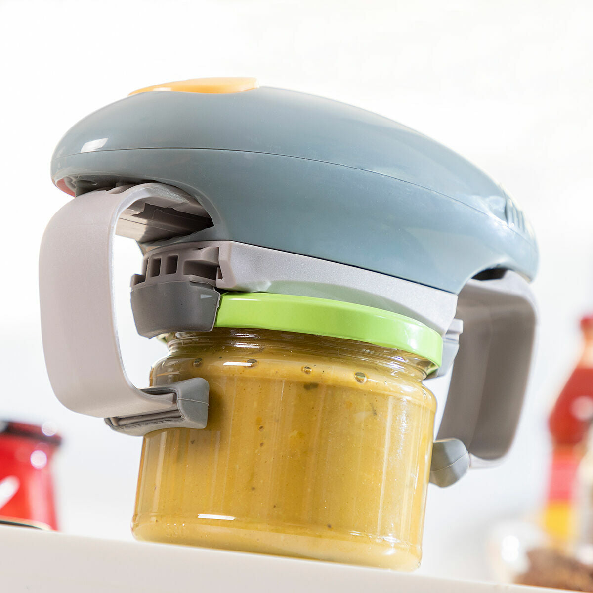 The Electric Jar Opener Lloucan InnovaGoods shown firmly gripping a jar with a green lid in a kitchen environment. This image enhances the perception of its strength and utility.