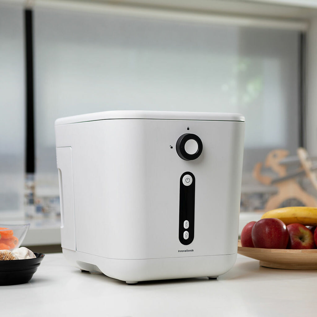 The composter is placed on a kitchen countertop with nearby fruits and vegetables. The background features a person.