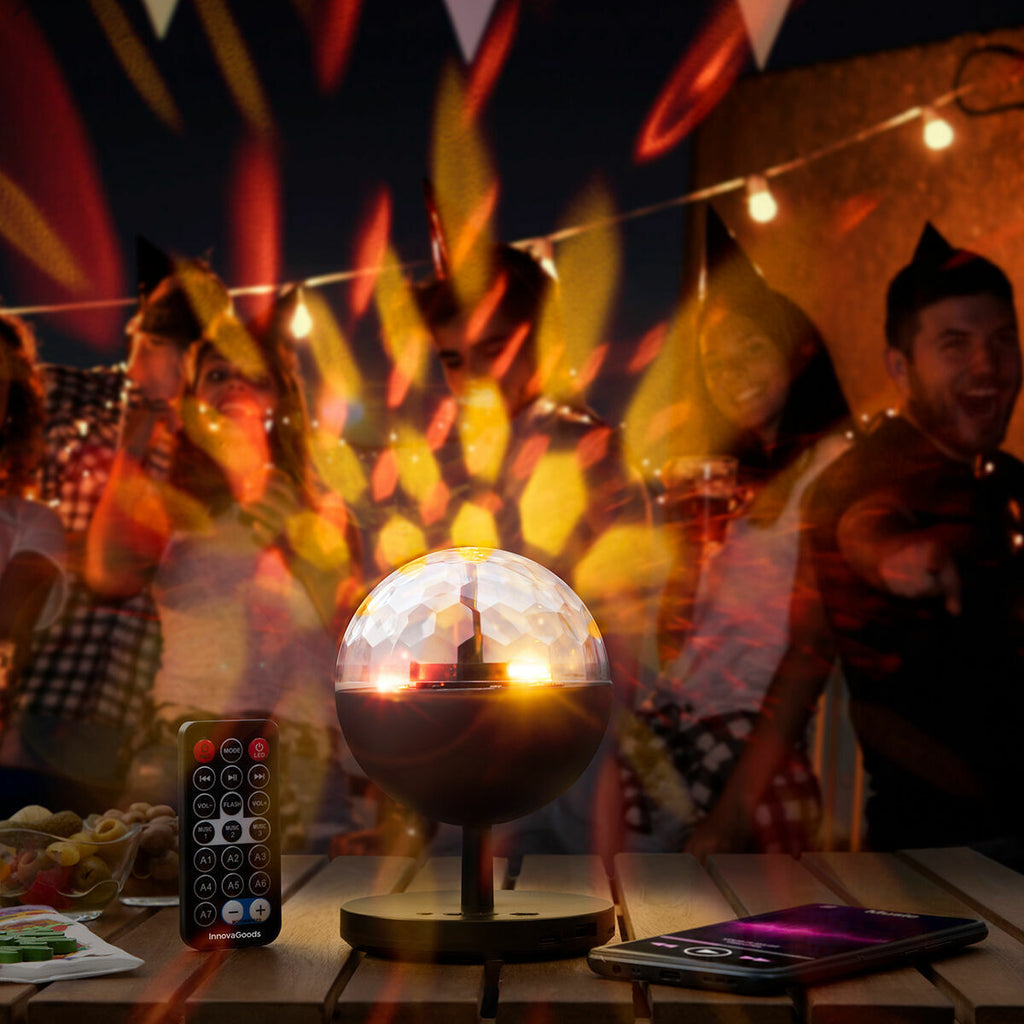 The image depicts the Rechargeable LED Projector with Speaker Istarlyt InnovaGoods on a table in a party setting, illuminating the area with colorful lights. The background shows people enjoying the party, enhancing its social atmosphere.