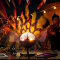 The image depicts the Rechargeable LED Projector with Speaker Istarlyt InnovaGoods on a table in a party setting, illuminating the area with colorful lights. The background shows people enjoying the party, enhancing its social atmosphere.