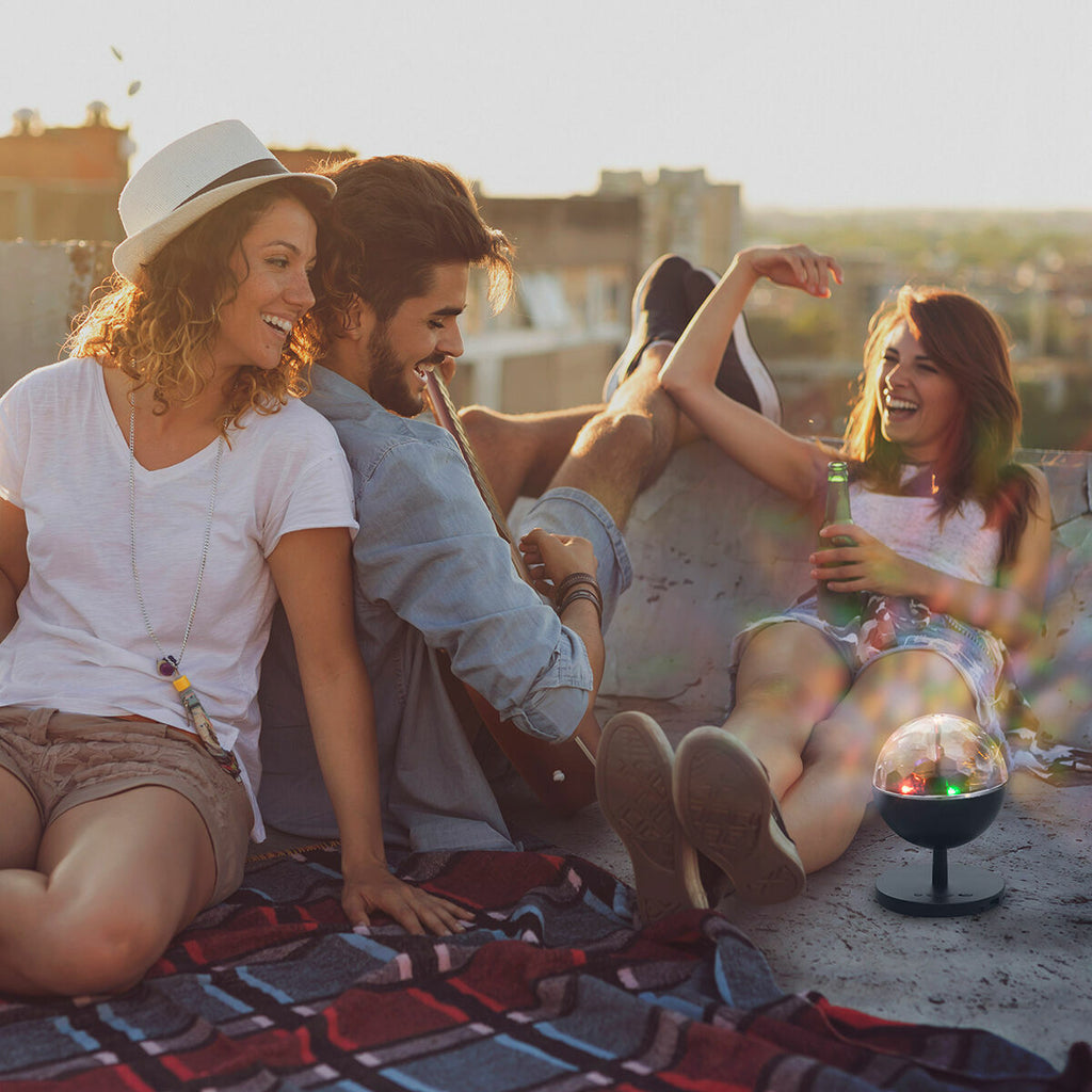 A lifestyle image featuring the Rechargeable LED Projector with Speaker Istarlyt InnovaGoods in a relaxed rooftop setting. People are enjoying the atmosphere with the device contributing to the ambience with its colorful lights.