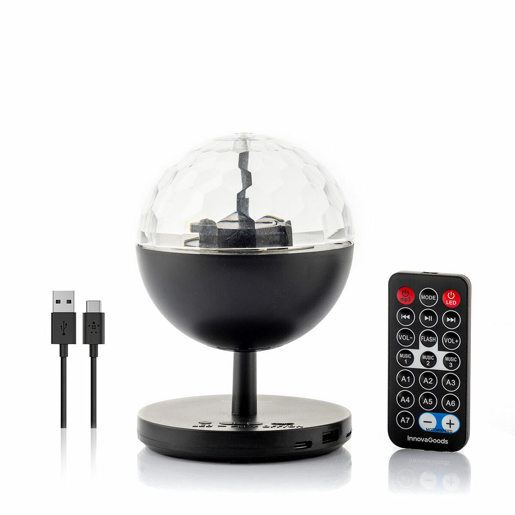 A detailed product shot showing the Rechargeable LED Projector with Speaker Istarlyt InnovaGoods along with its remote control and USB cables. The projector has a spherical design with a faceted dome.