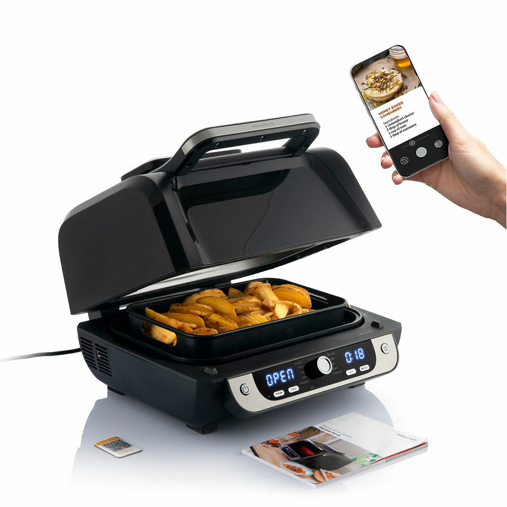 The air fryer in use, with food inside and a hand holding a smartphone displaying a recipe.