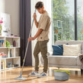Person using a grey and yellow self-cleaning spin mop with separation bucket in a well-lit room with wooden floors.