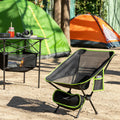 A lifestyle image showing the camping chair set up at a campsite with a table and two tents in the background. The chair is black with lime green accents and includes a mesh seat and storage pockets. The setting conveys a comfortable camping experience.