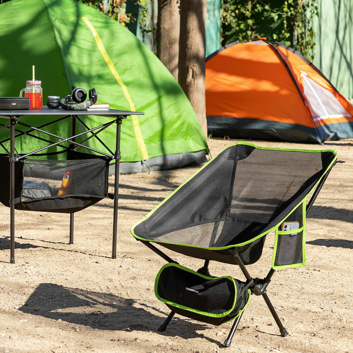 A lifestyle image showing the camping chair set up at a campsite with a table and two tents in the background. The chair is black with lime green accents and includes a mesh seat and storage pockets. The setting conveys a comfortable camping experience.