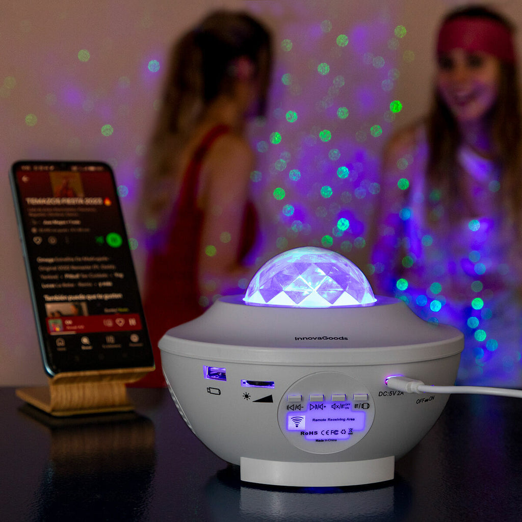 The LED Star Projector with Speaker is displayed in a party setting, projecting green and blue lights. It is placed on a table beside a smartphone. Two people are visible in the blurred background, enjoying the festive atmosphere created by the light patterns.