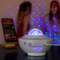 The LED Star Projector with Speaker is displayed in a party setting, projecting green and blue lights. It is placed on a table beside a smartphone. Two people are visible in the blurred background, enjoying the festive atmosphere created by the light patterns.