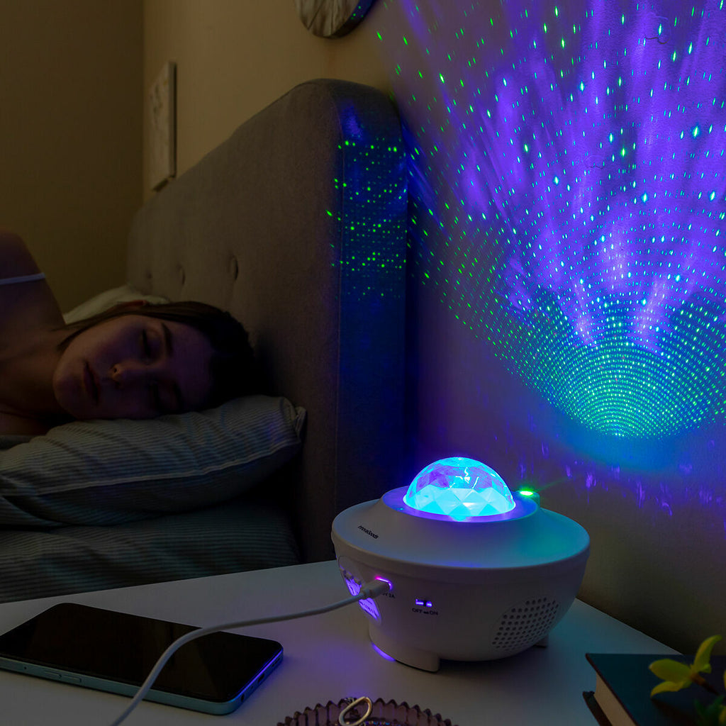 The projector in a calm bedroom setting, projecting star patterns on the wall while a person sleeps. The device is on a bedside table next to a smartphone.