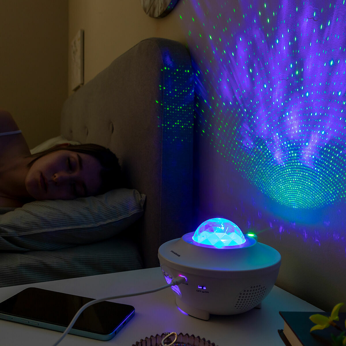 The projector in a calm bedroom setting, projecting star patterns on the wall while a person sleeps. The device is on a bedside table next to a smartphone.