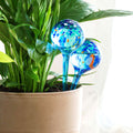Two watering globes inserted into a plant pot, surrounded by lush green leaves, highlighting their function in an indoor setting.