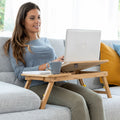 A woman using a bamboo folding side table on a couch, holding a laptop and a coffee cup on the table. The setting is in a living room with a grey couch and colorful cushions.