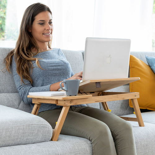 A woman using a bamboo folding side table on a couch, holding a laptop and a coffee cup on the table. The setting is in a living room with a grey couch and colorful cushions.