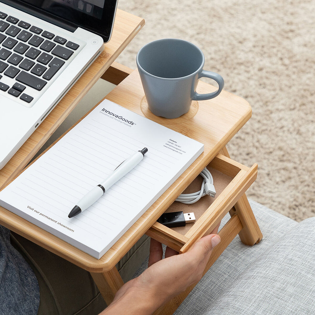 Close-up showing the drawer of the bamboo table, storing accessories like cables and pens, with a cup and notepad on the surface.