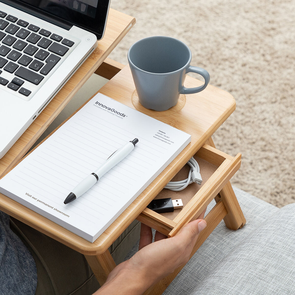 Close-up showing the drawer of the bamboo table, storing accessories like cables and pens, with a cup and notepad on the surface.