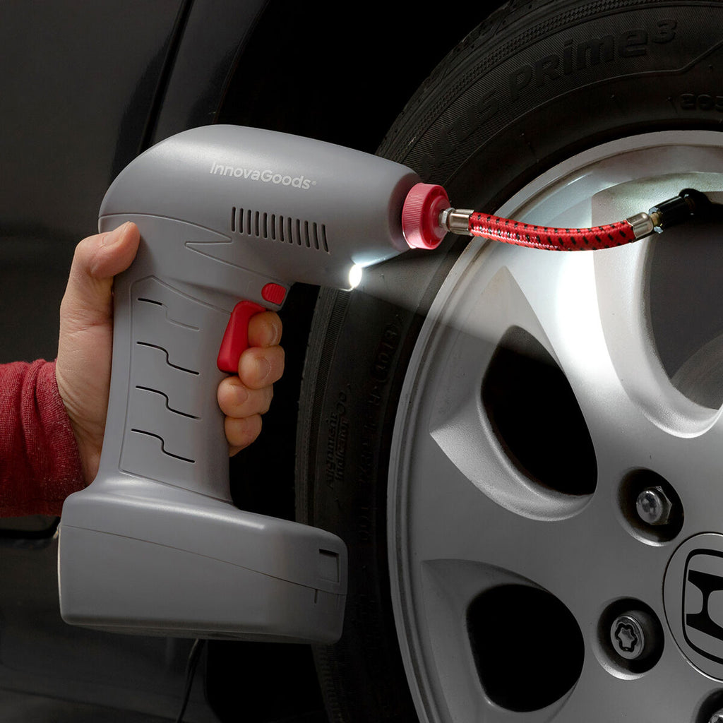 A gray InnovaGoods portable air compressor with a red trigger and a red nozzle. The device is being used to inflate a car tire, with an LED light illuminating the area.
