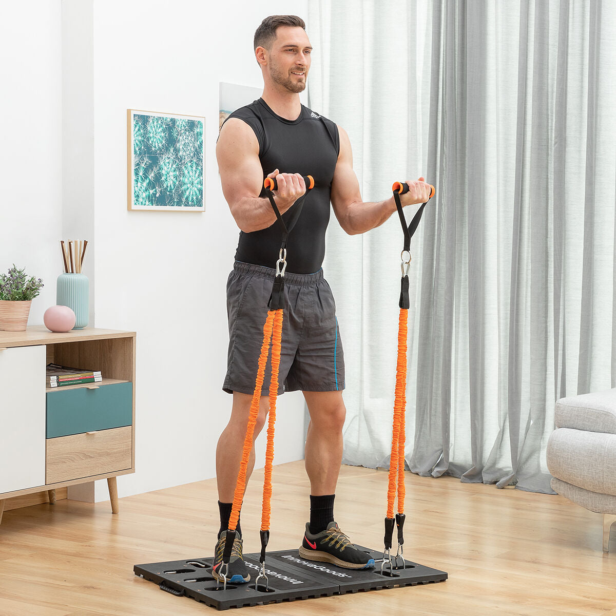 A man uses the training system indoors, standing on the platform, exercising with orange bands and handles. The setting is a well-lit living room.
