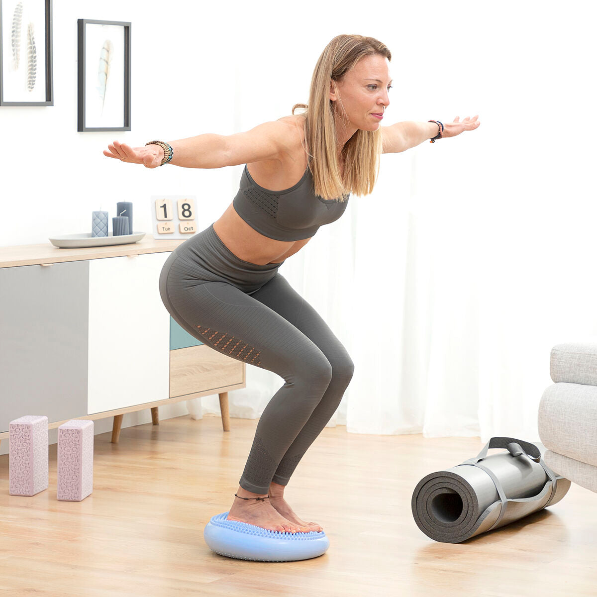 A woman in athletic wear using the balance cushion for squats in a home setting. The cushion is light blue with a textured surface.