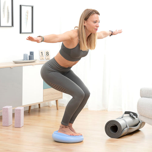 A woman in athletic wear using the balance cushion for squats in a home setting. The cushion is light blue with a textured surface.