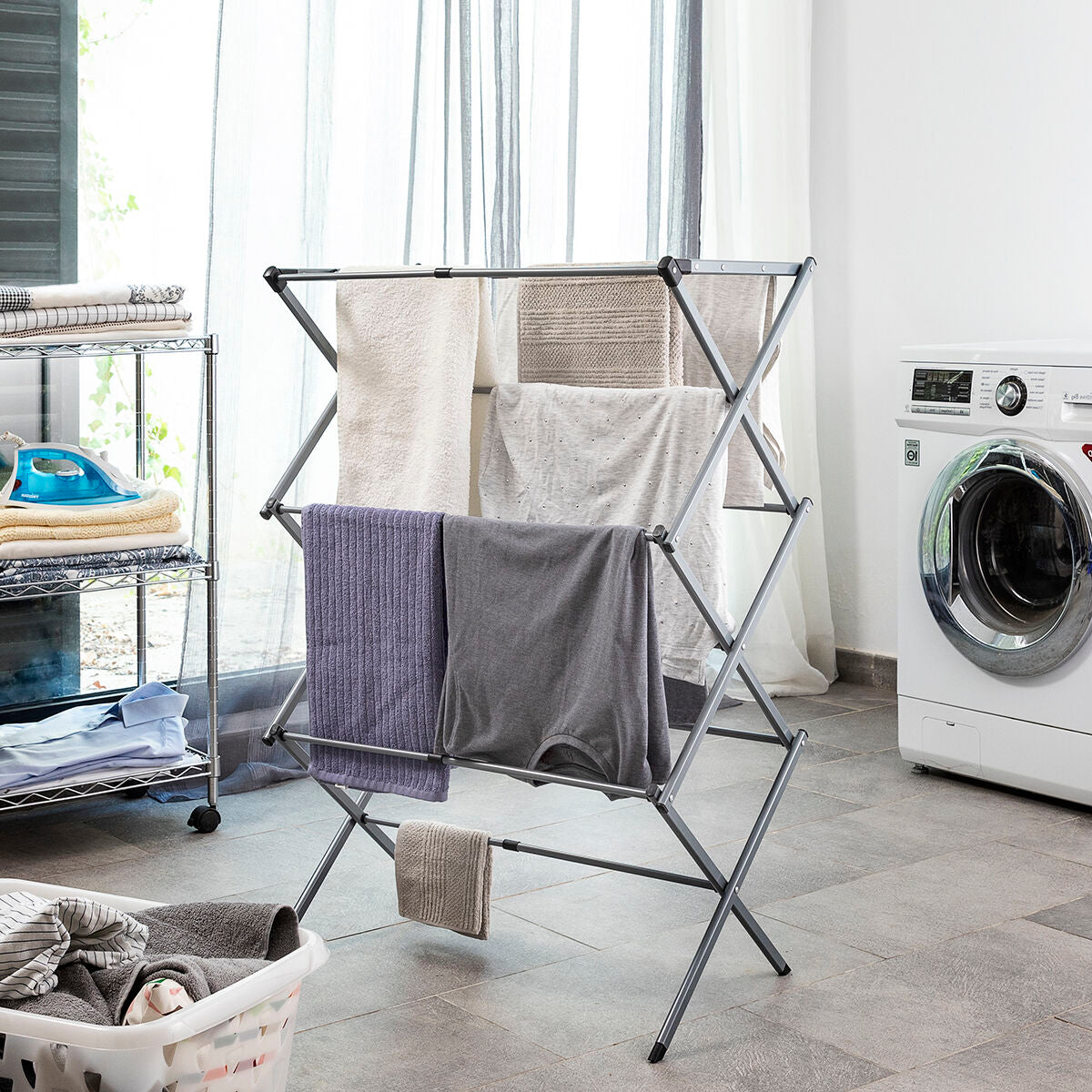 A metal clothes dryer standing in a laundry room. The dryer is open with clothes hanging on it, placed beside a washing machine. The floor and subtle window light add to the context.