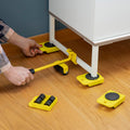 A person using the yellow 'HeavEasy Lifting and Moving Tool' with a lever to place a wheeled pad under a piece of furniture on a wooden floor.