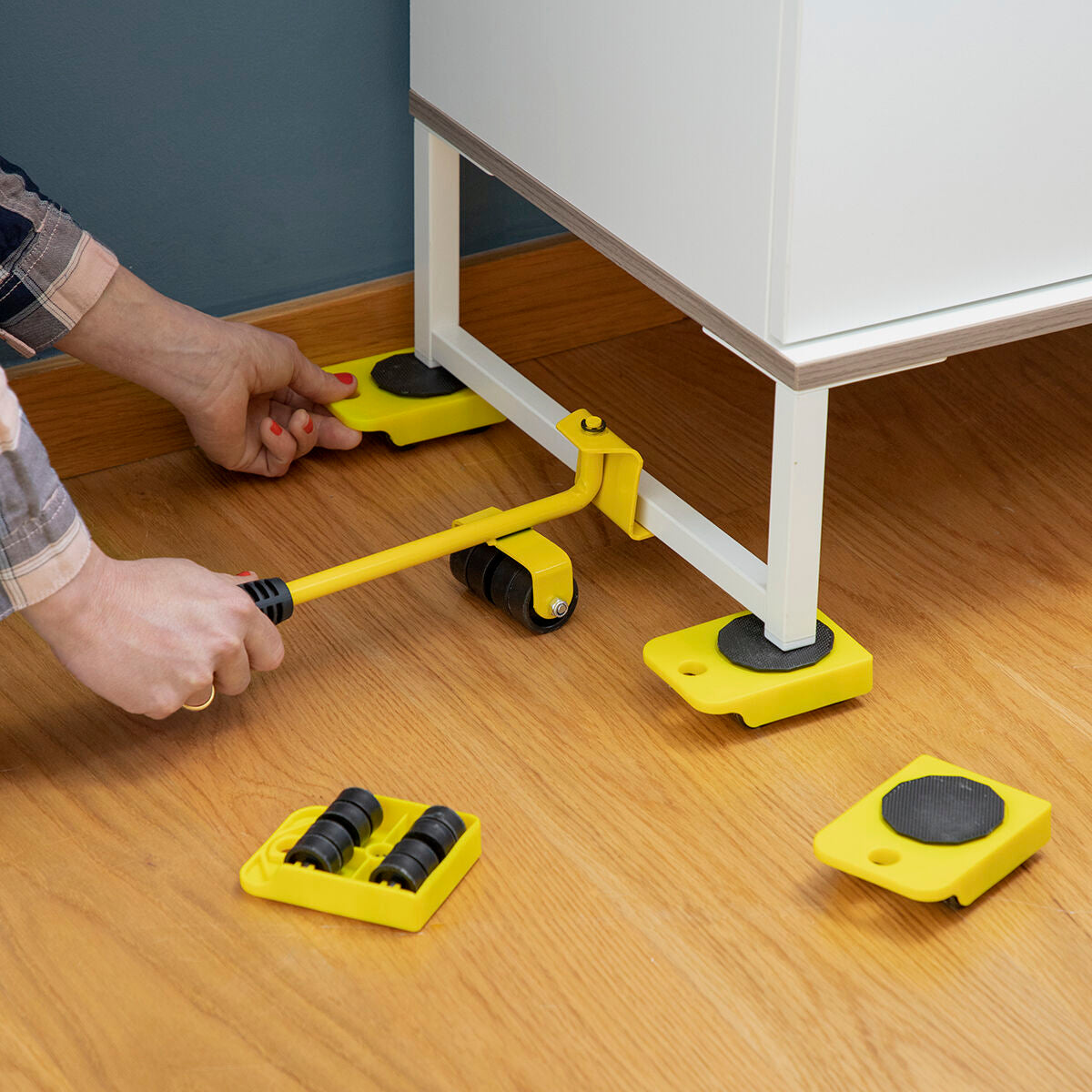 A person using the yellow 'HeavEasy Lifting and Moving Tool' with a lever to place a wheeled pad under a piece of furniture on a wooden floor.