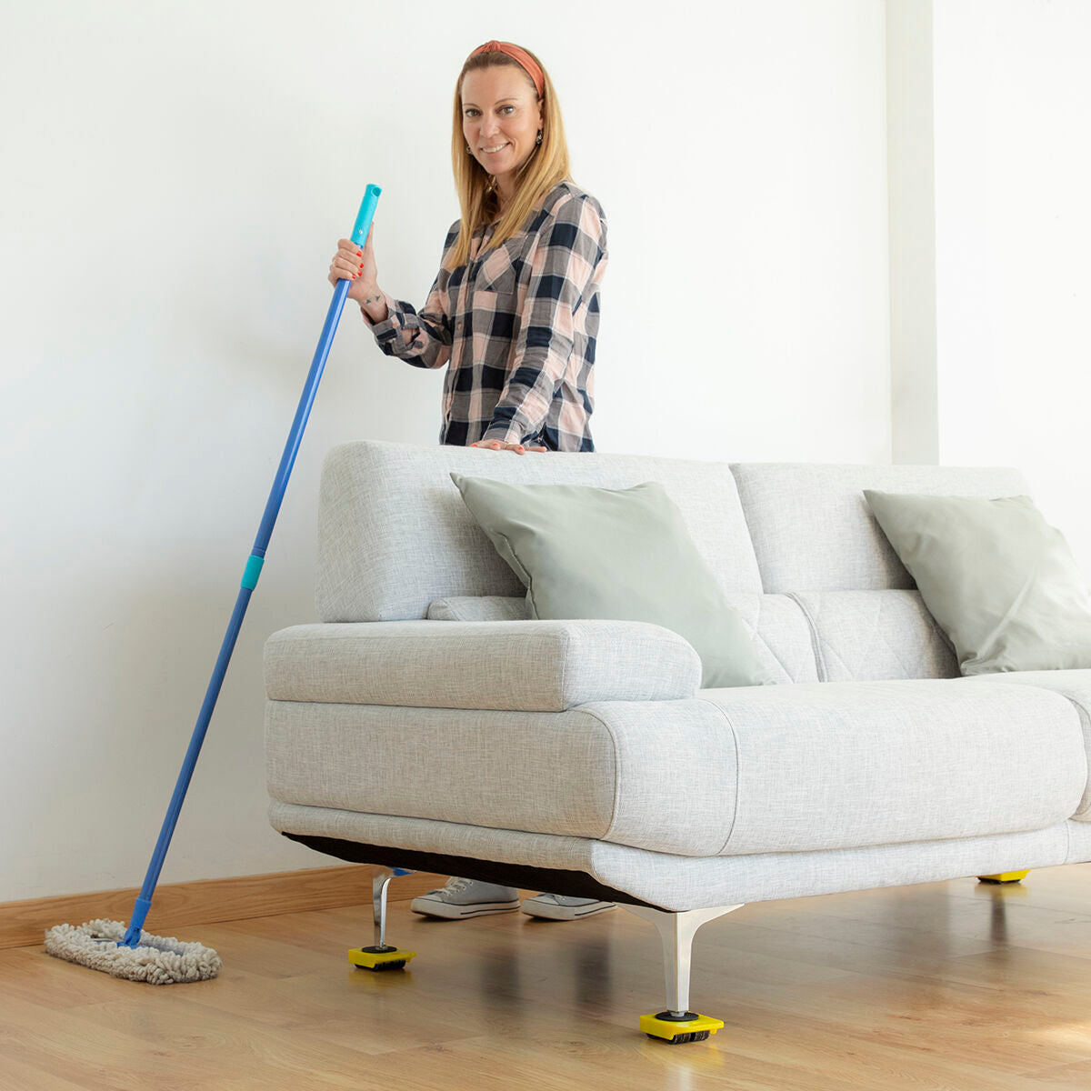 A person standing in a room with a couch on yellow pads from the 'HeavEasy Lifting and Moving Tool' as they hold a mop.