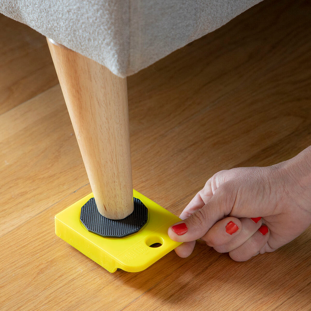 A close-up of a person placing a yellow wheeled pad under a couch leg using the 'HeavEasy Lifting and Moving Tool'.