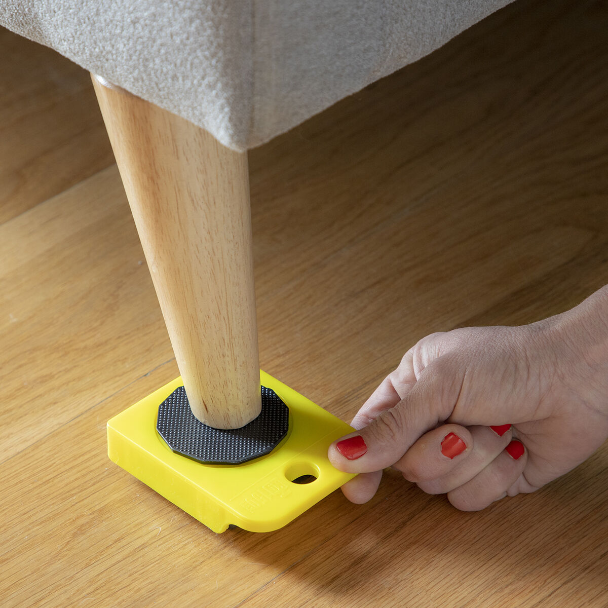 A close-up of a person placing a yellow wheeled pad under a couch leg using the 'HeavEasy Lifting and Moving Tool'.