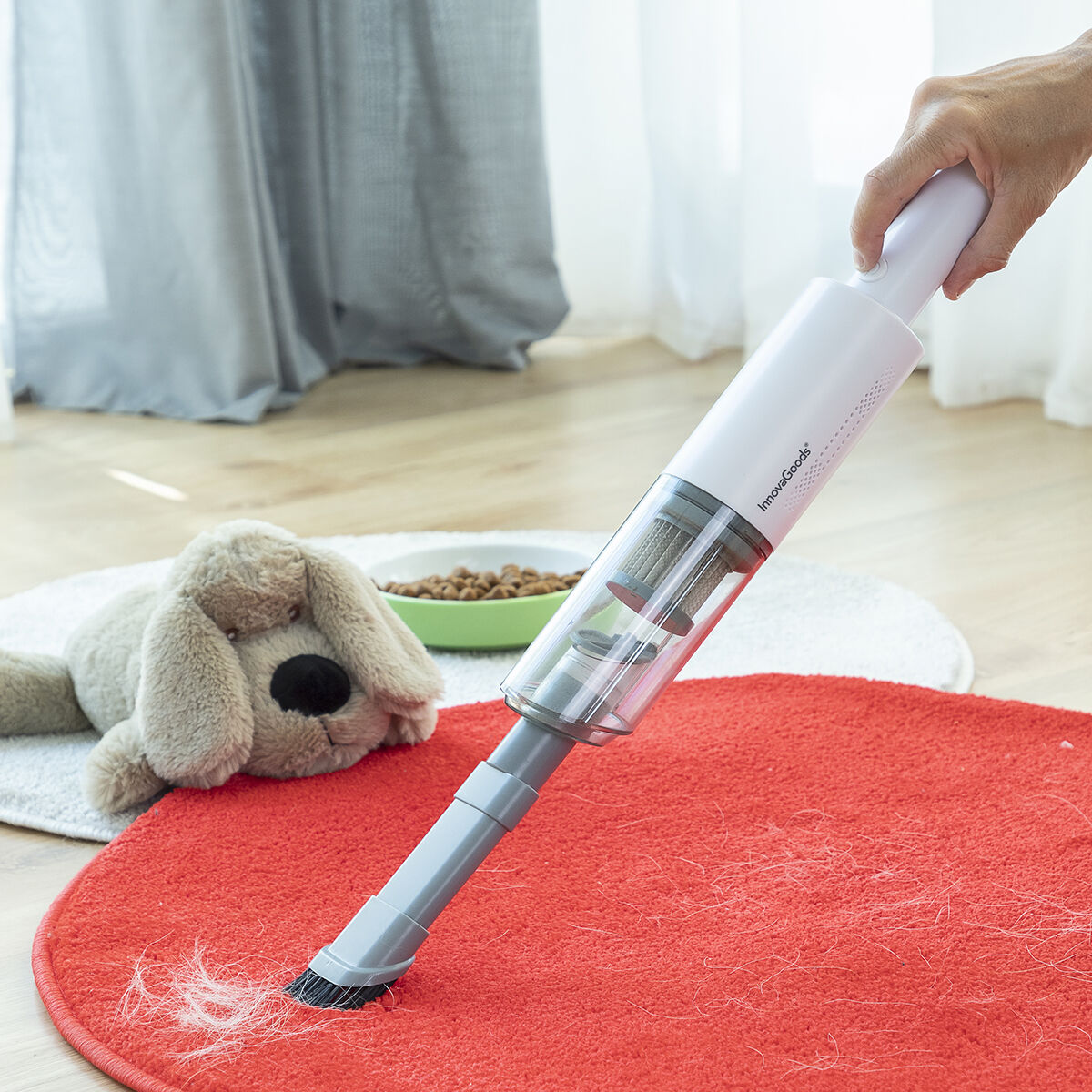 The handheld vacuum cleaner is being used to clean pet hair off a red carpet. The vacuum is white with a transparent section, showing the filter inside. There's a stuffed animal and pet food bowl in the background, indicating a pet-friendly environment. The hand holding the device is visible, demonstrating its lightweight and ergonomic design.