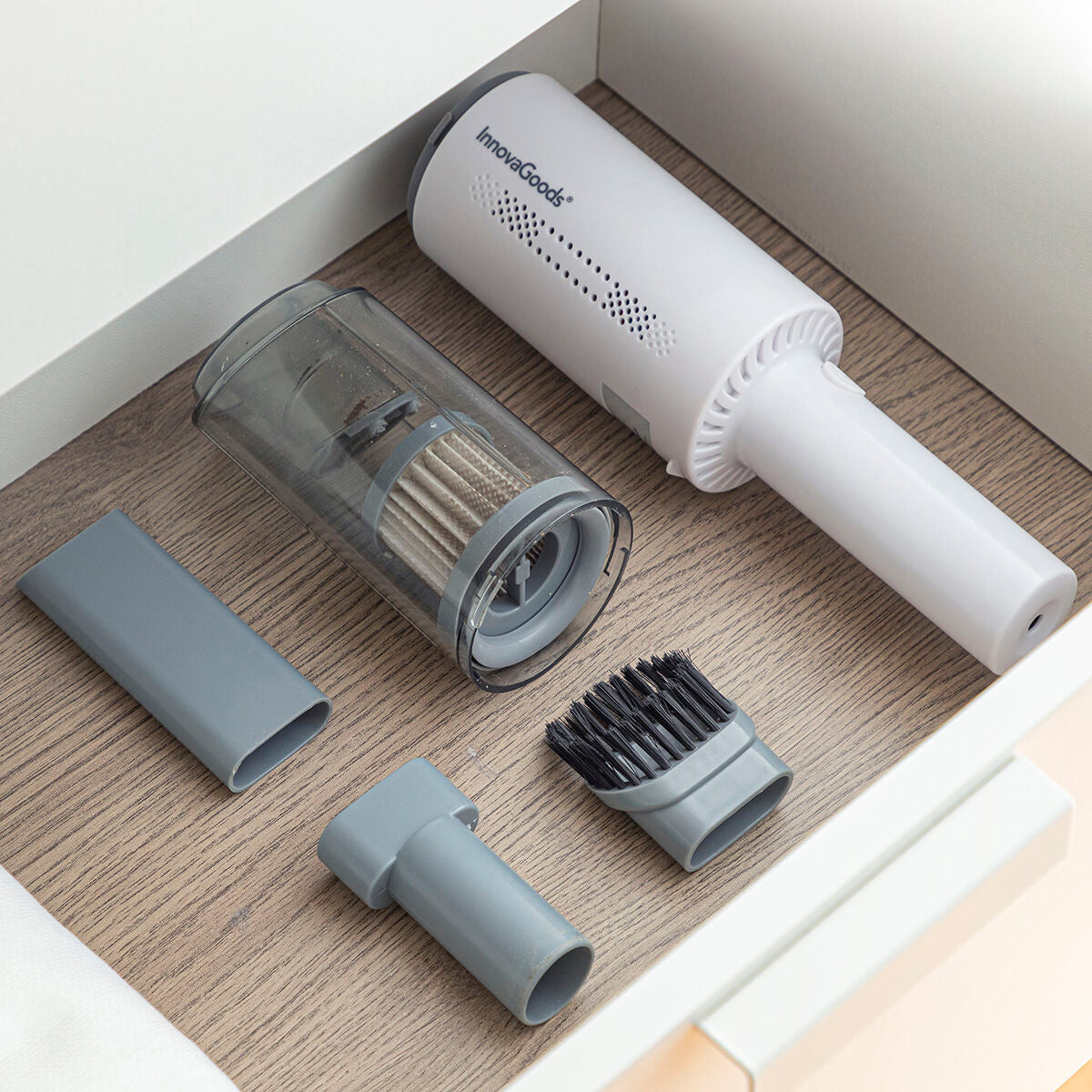 Shows the handheld vacuum and its accessories neatly arranged in a drawer, demonstrating its compact storage capability. The vacuum's parts, including the gray attachments, are organized on a wooden surface.