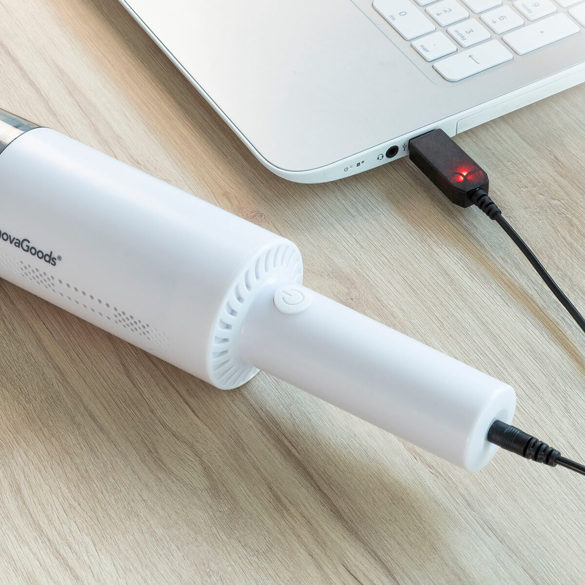 The vacuum cleaner plugged into a laptop via a USB charging cable, demonstrating its rechargeable feature. The device is laid flat on a wooden surface, emphasizing its practicality and convenience in charging.
