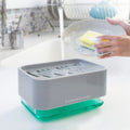 A lifestyle image of the InnovaGoods Pushoap 2-in-1 Soap Dispenser in use in a kitchen setting. A person is using a sponge on a plate next to the dispenser, emphasizing its practical use. The dispenser is gray with a transparent bottom section filled with green soap.