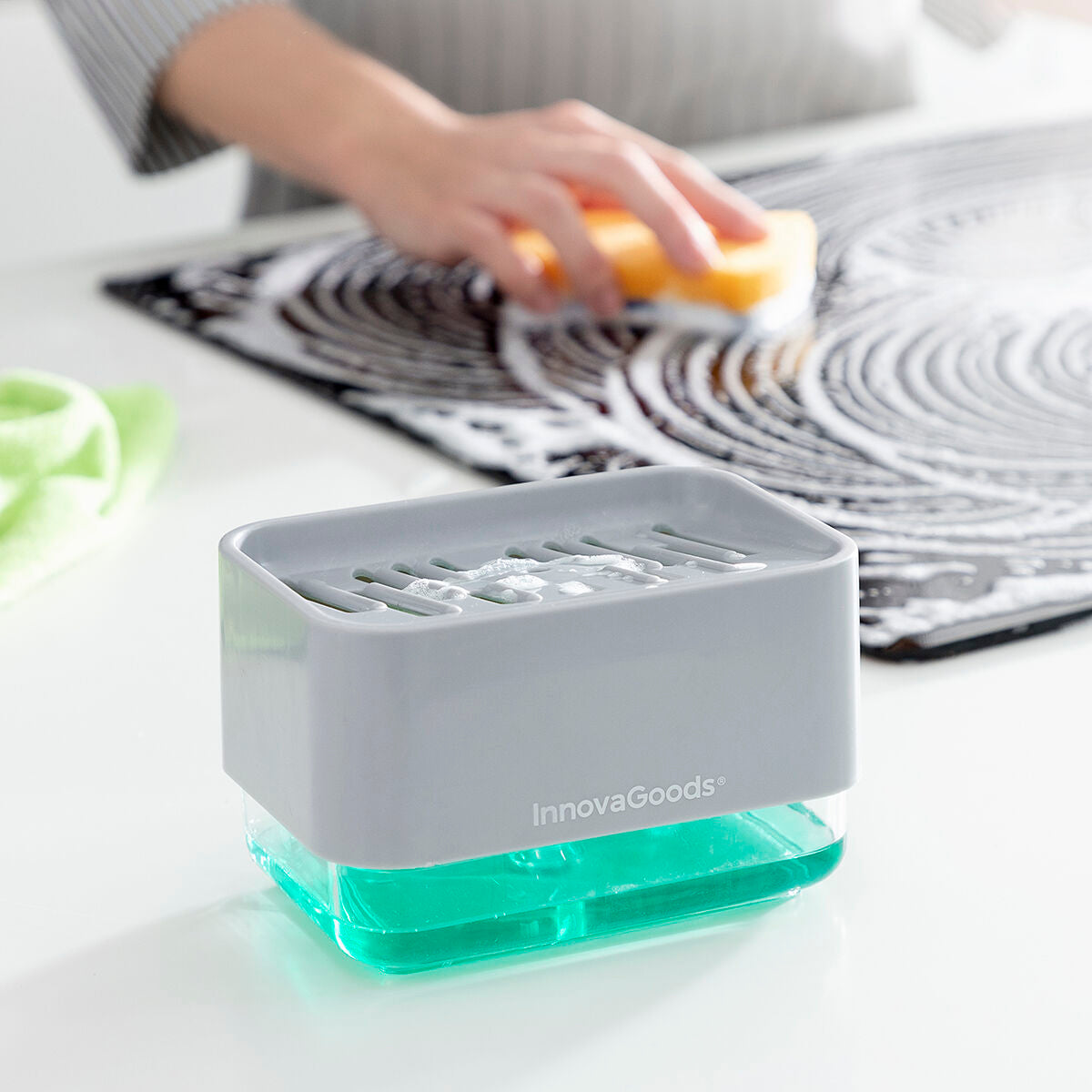 The InnovaGoods Pushoap 2-in-1 Soap Dispenser in use on a countertop with someone cleaning a surface with soap. The gray and transparent design is visible, with a focus on the dispenser dispensing soap efficiently.