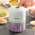 A lifestyle image showing the InnovaGoods mini chopper being used to chop purple onions. A hand is pressing the button on top, with various chopped ingredients surrounding the device.