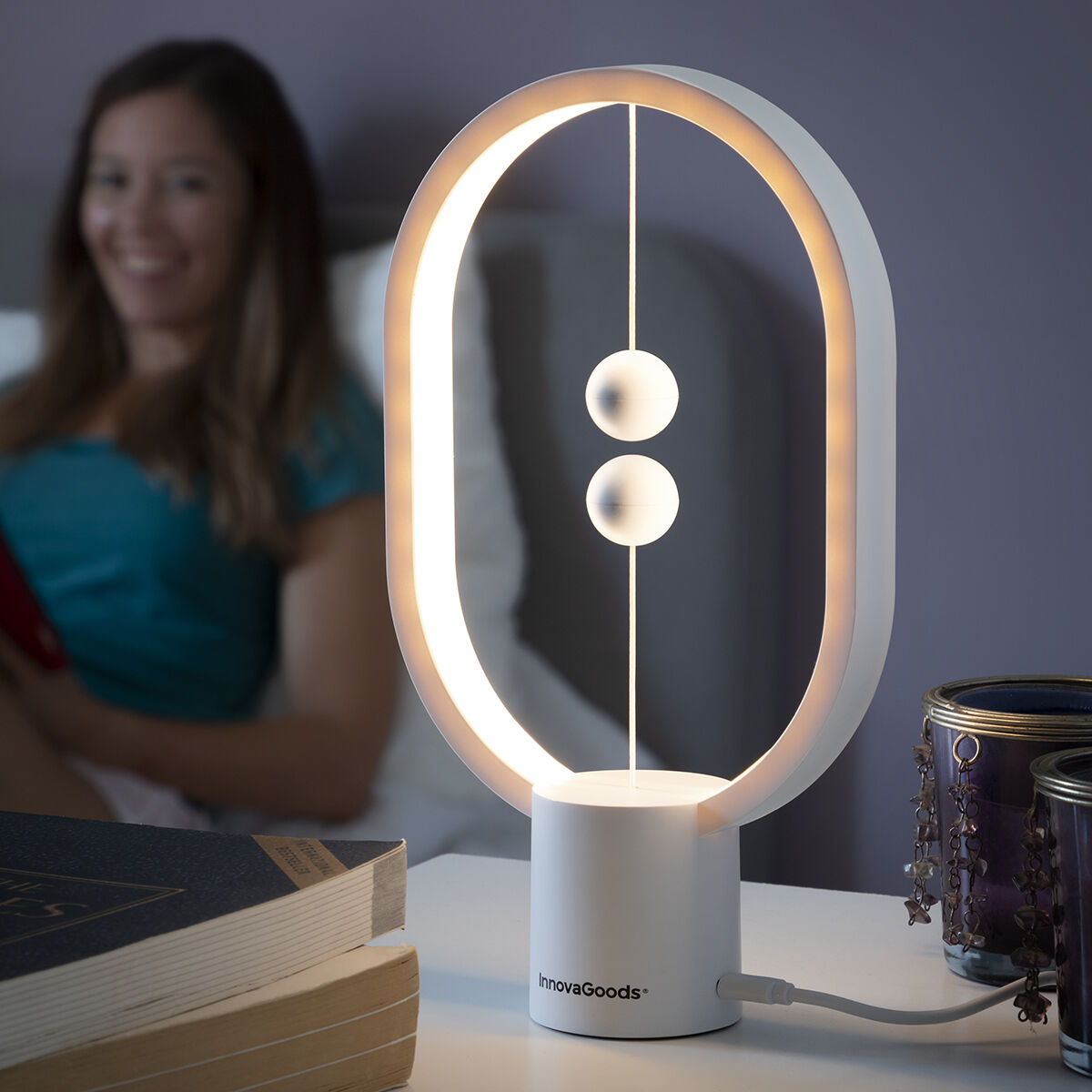 Lifestyle image of the InnovaGoods Balance Lamp with Magnetic Switch. The lamp is on a bedside table, emitting a warm light, with a blurred person in the background on a bed, suggesting a cozy and relaxing setting. The lamp's magnetic spheres are visible, indicating its unique switch mechanism. Books and decorative items surround the lamp.