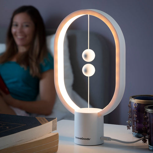 Lifestyle image of the InnovaGoods Balance Lamp with Magnetic Switch. The lamp is on a bedside table, emitting a warm light, with a blurred person in the background on a bed, suggesting a cozy and relaxing setting. The lamp's magnetic spheres are visible, indicating its unique switch mechanism. Books and decorative items surround the lamp.