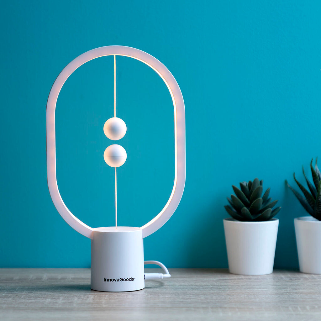 Lifestyle image of the InnovaGoods Balance Lamp on a wooden surface next to decorative plants. The lamp is lit, providing ambient lighting in a modern, stylish setting.