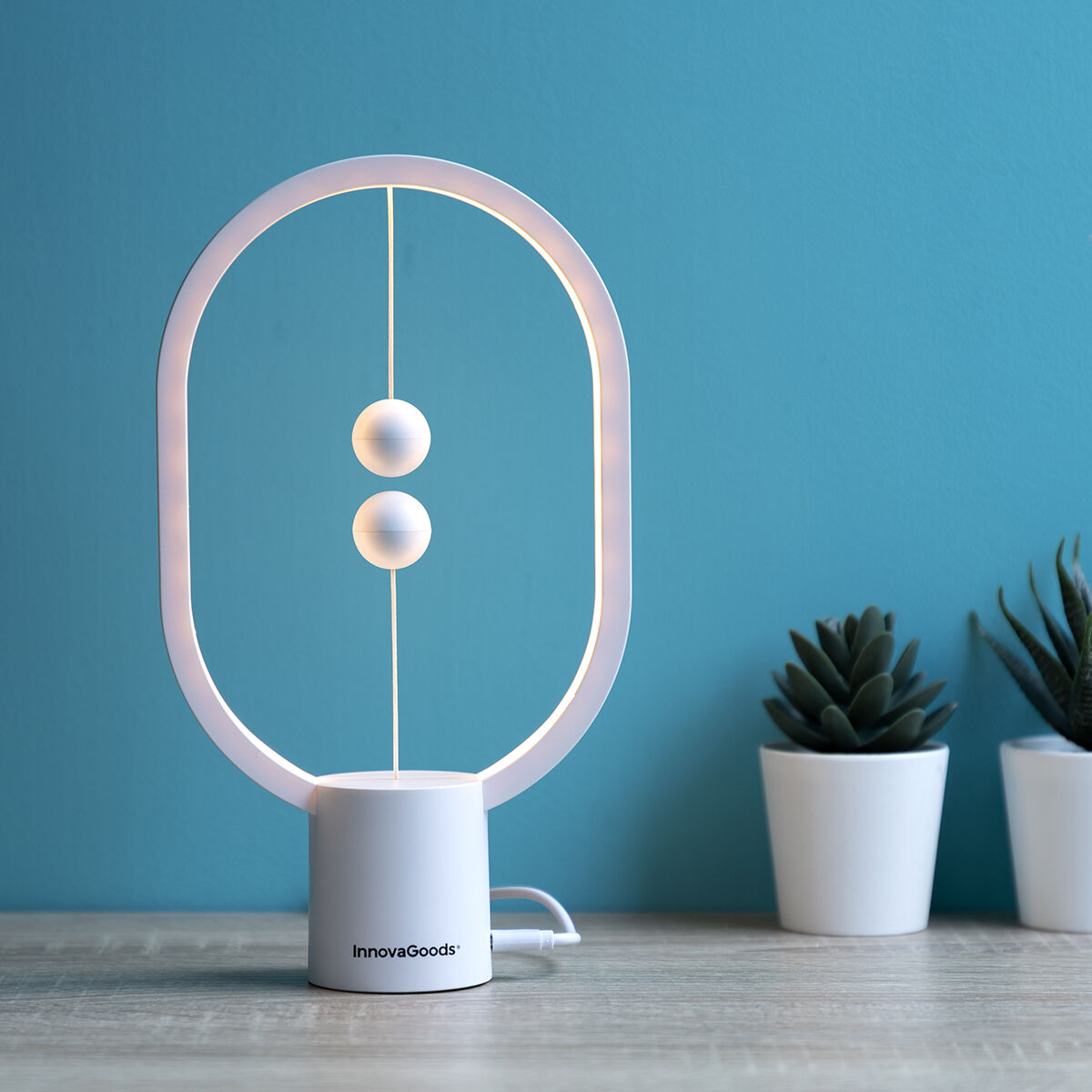 Lifestyle image of the InnovaGoods Balance Lamp on a wooden surface next to decorative plants. The lamp is lit, providing ambient lighting in a modern, stylish setting.